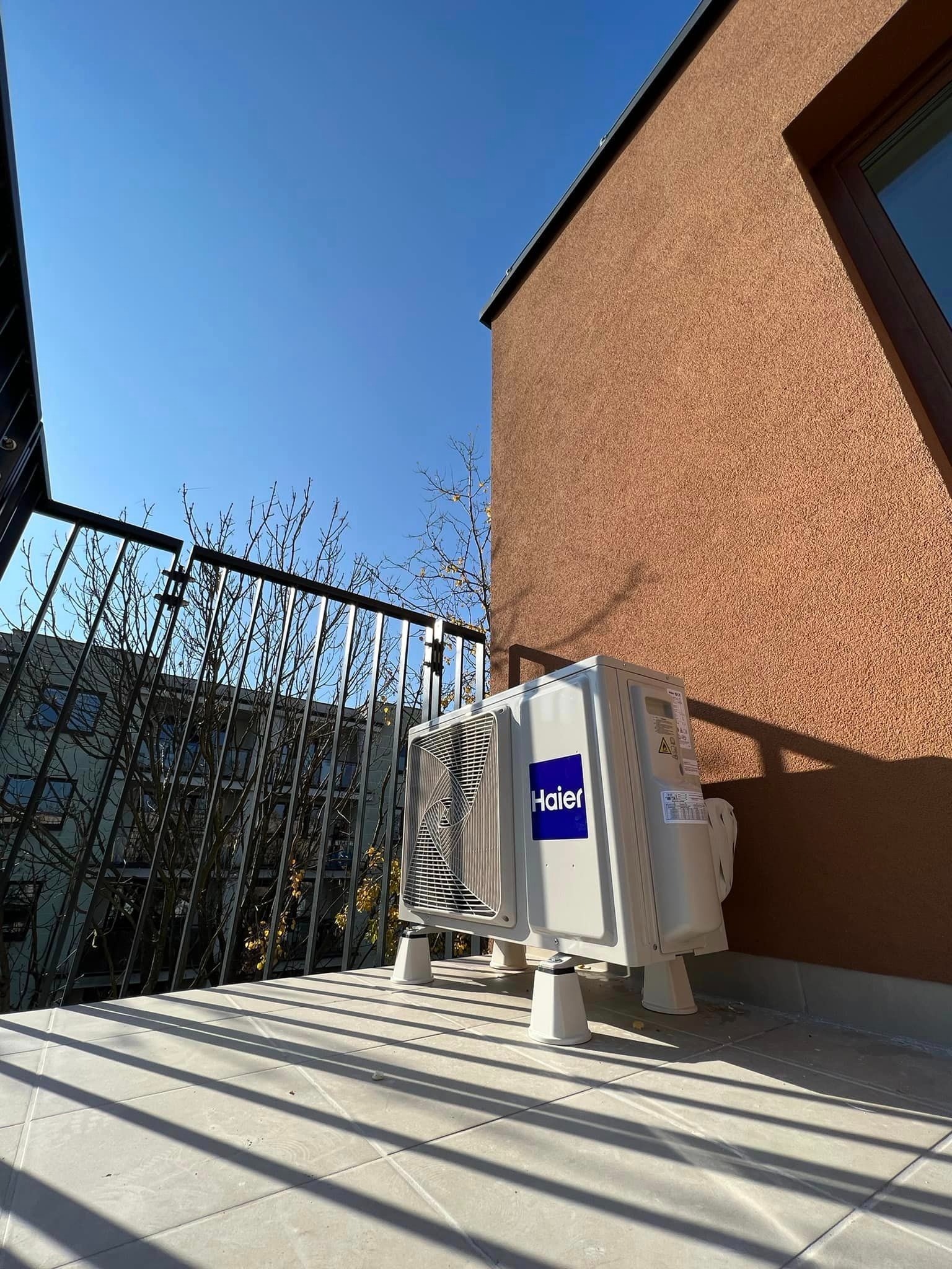 Zewnętrzna jednostka klimatyzacji Haier na balkonie, zamontowana na podwyższeniach, widok na błękitne niebo i budynek z brązową elewacją.