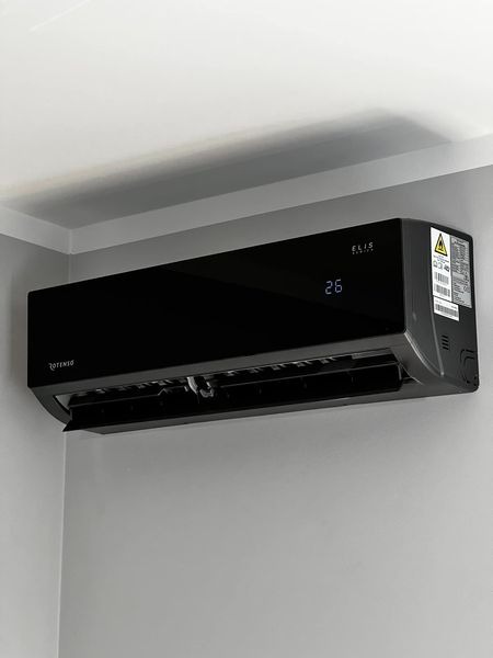 Czarny klimatyzator ścienny marki Rotenso Elis z wyświetlaczem temperatury 26 stopni Celsjusza, zamontowany w rogu pomieszczenia przy białym suficie.