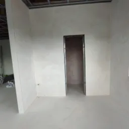 Surowe wnętrze w trakcie prac wykończeniowych: ściany pokryte szarą warstwą, widoczna metalowa konstrukcja sufitu podwieszanego oraz surowy otwór drzwiowy z widocznymi bloczkami betonowymi wewnątrz.