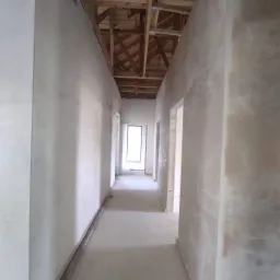 Korytarz w stanie surowym, ściany pokryte wstępną warstwą tynku, widoczne drewniane elementy konstrukcji dachu.