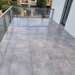 WoodworkGroup.pl - Nowoczesny balkon z szarymi płytkami ceramicznymi, szklaną balustradą i widokiem na teren budowy w tle, uzupełniony logo firmy Woodwork Group.pl na ścianie.