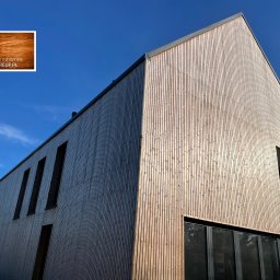 WoodworkGroup.pl - Nowoczesny budynek z elewacją z jasnego drewna o pionowym układzie desek, z wyraźnym logo firmy w rogu, na tle błękitnego nieba.