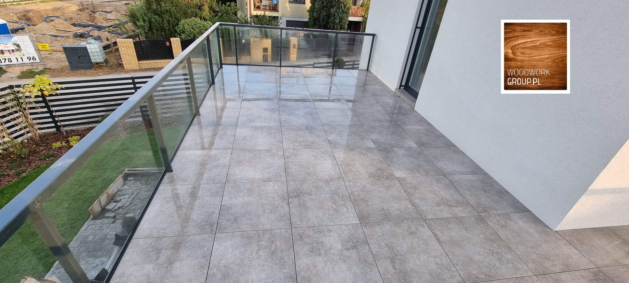 Nowoczesny balkon z szarymi płytkami ceramicznymi, szklaną balustradą i widokiem na teren budowy w tle, uzupełniony logo firmy Woodwork Group.pl na ścianie.