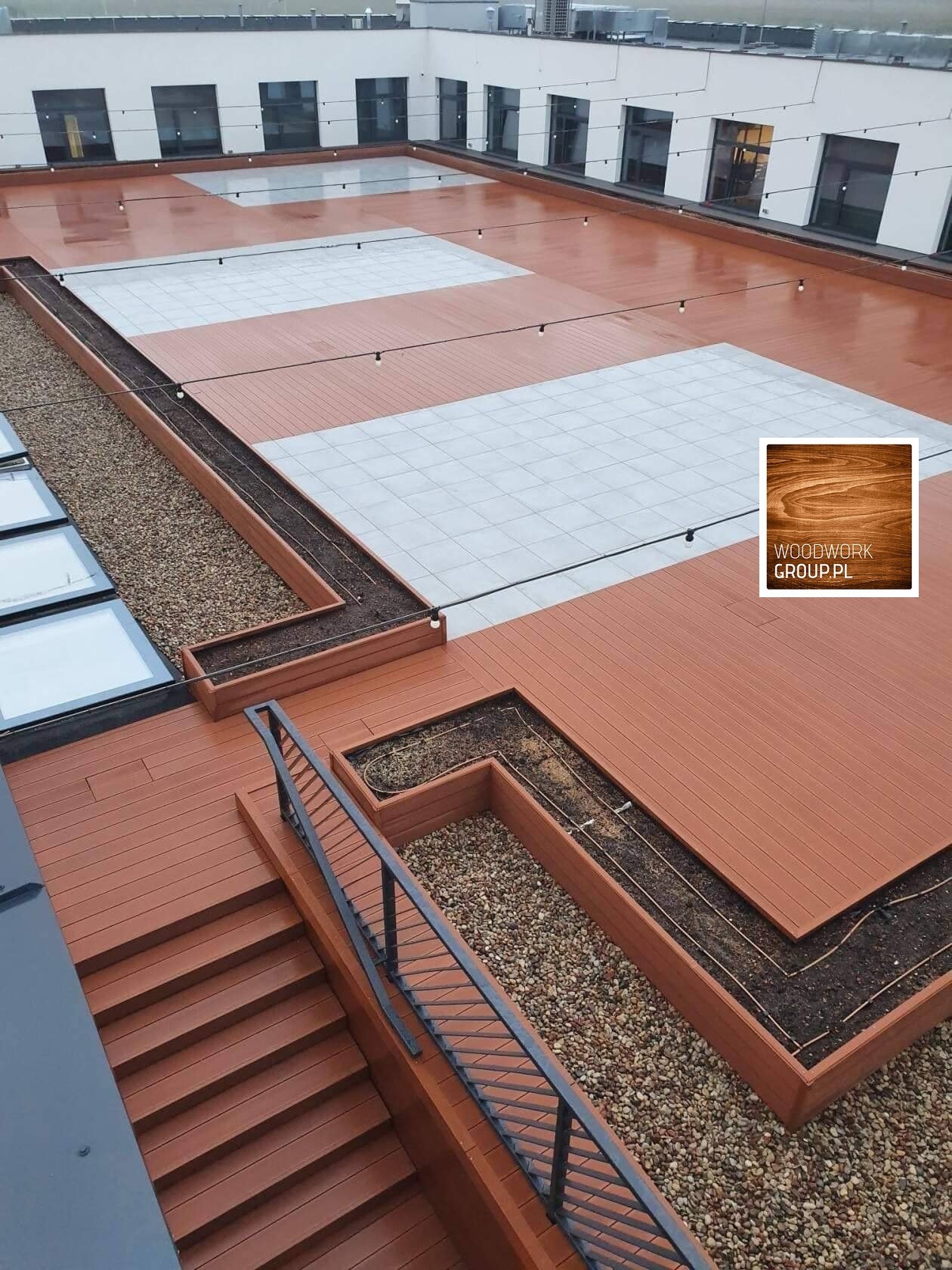 Rozległy taras na dachu budynku z brązowych desek, urozmaicony fragmentami z jasnych płyt i wbudowanymi rabatami z kamieniami, oświetlony girlandami żarówek, widok z góry z balustradą schodów.