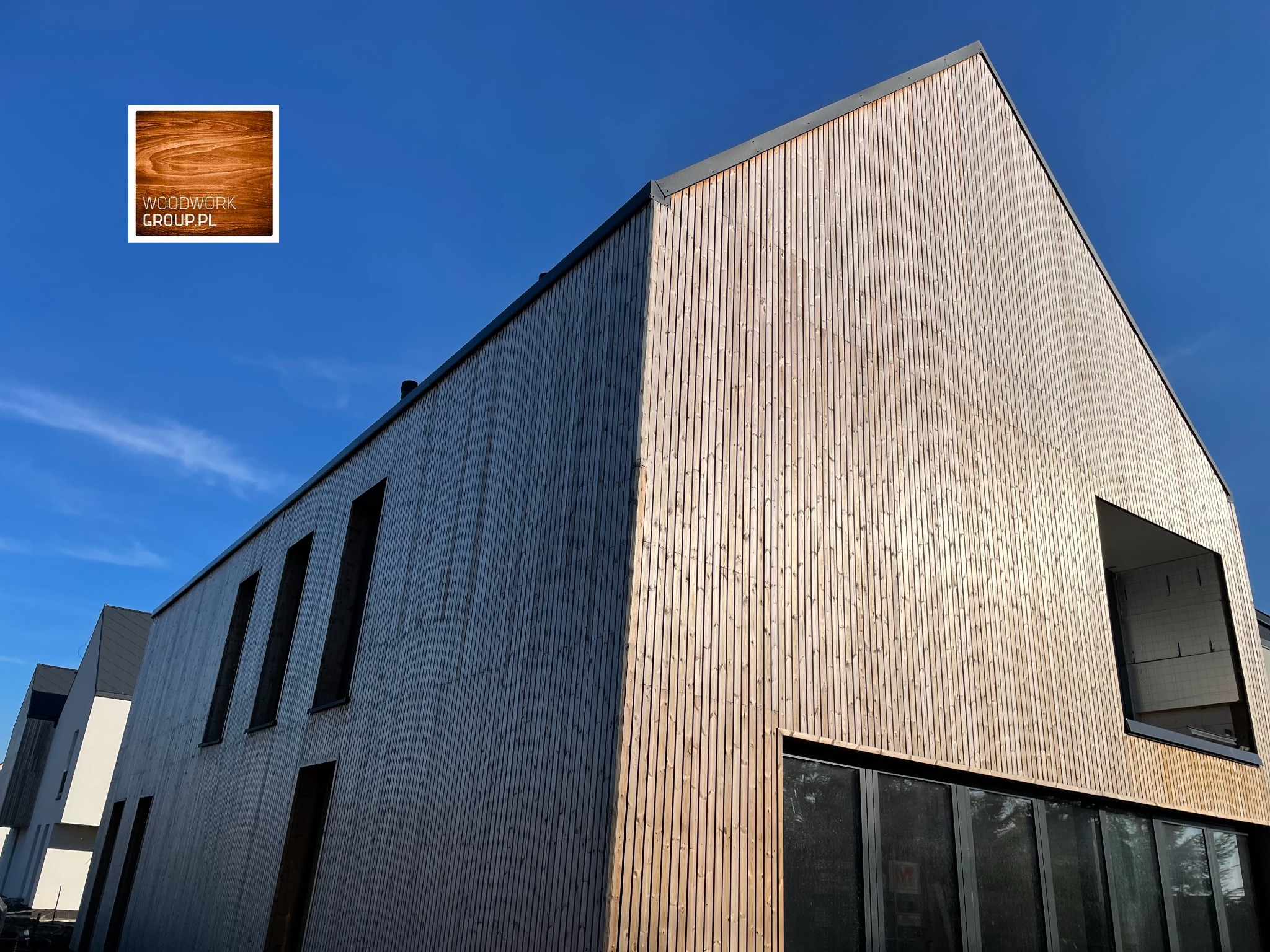Nowoczesny budynek z elewacją z jasnego drewna o pionowym układzie desek, z wyraźnym logo firmy w rogu, na tle błękitnego nieba.