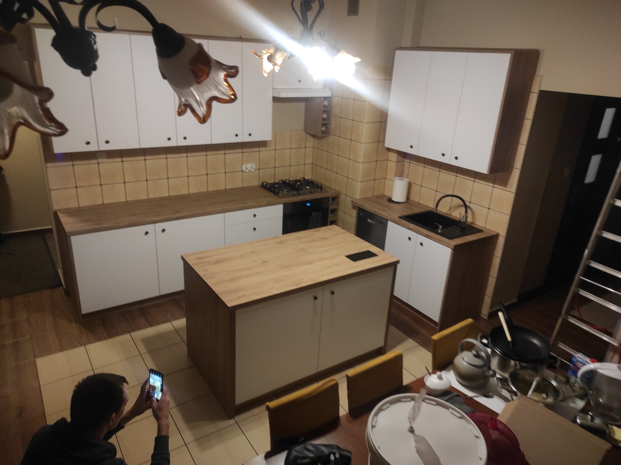 Wnętrze kuchni z wyspą, widziane z perspektywy osoby robiącej zdjęcie telefonem. Widoczne białe szafki z drewnianymi blatami, kuchenka, zlew oraz elementy wyposażenia.