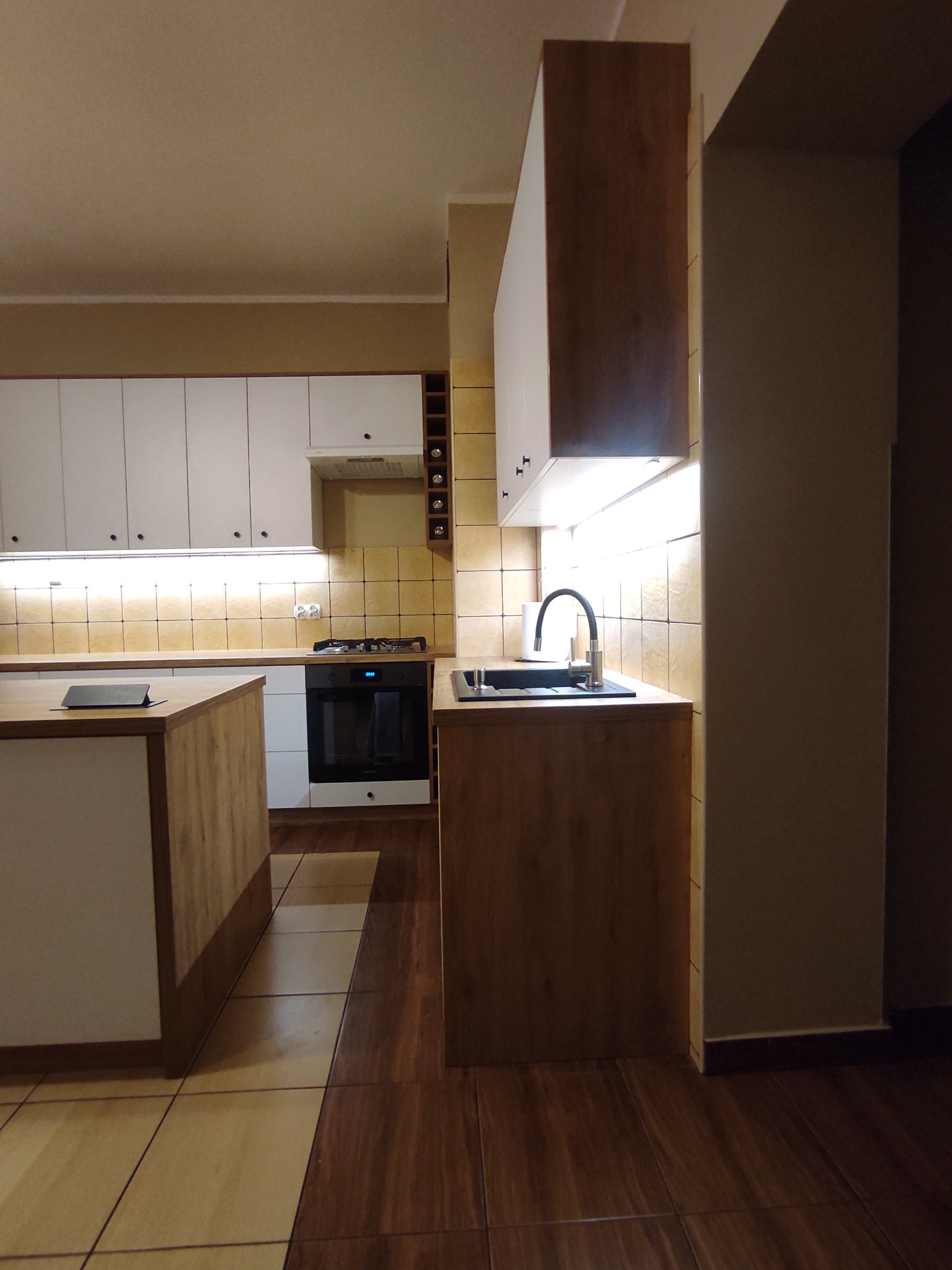 Wykończona kuchnia z białymi szafkami górnymi i dolnymi w odcieniach drewna, oświetleniem LED pod szafkami, brązową podłogą i płytkami na ścianie.