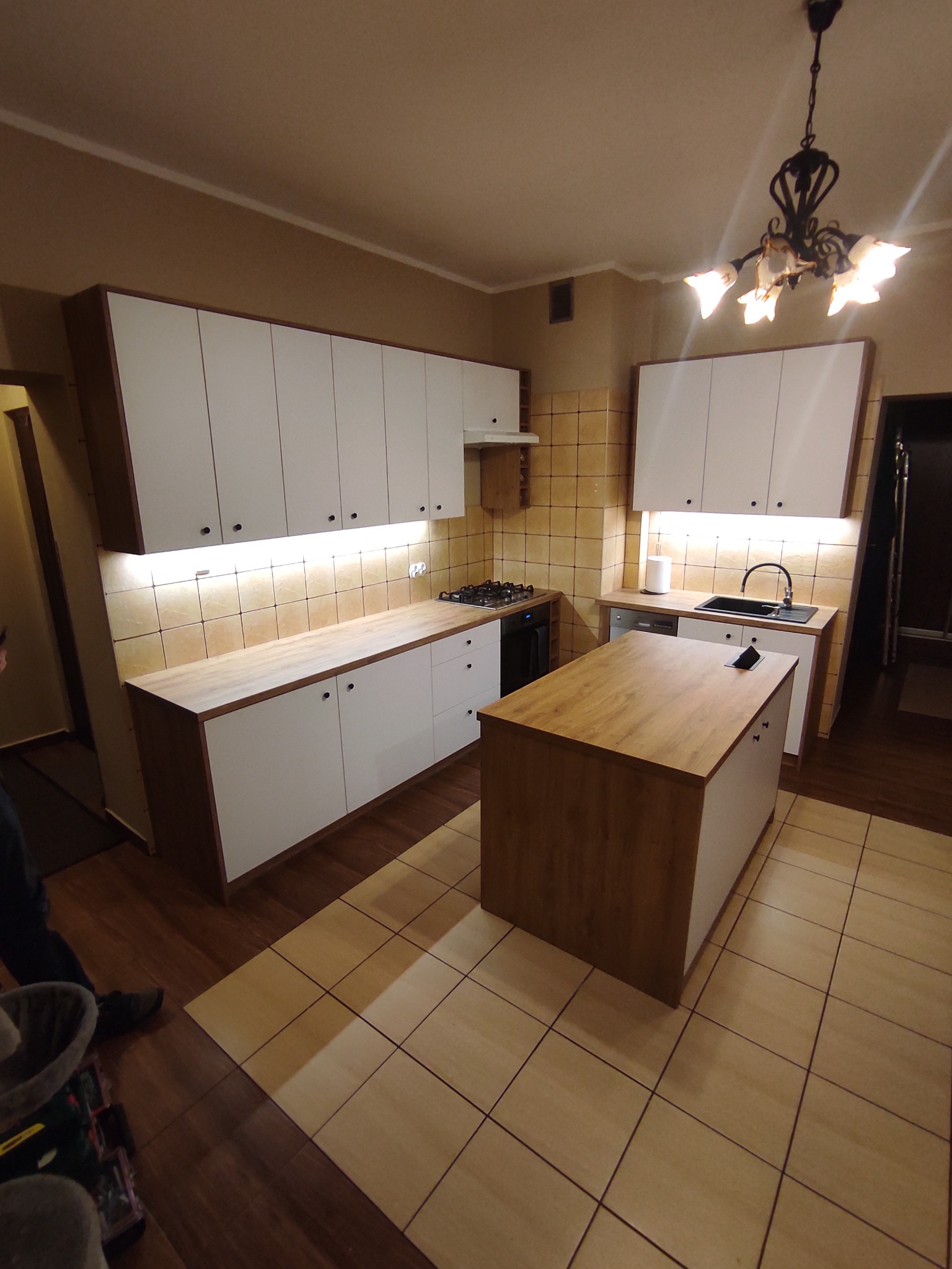 Nowoczesna kuchnia z białymi szafkami i drewnianymi blatami, oświetleniem LED podszafkowym i wyspą kuchenną na tle płytek w odcieniach beżu.