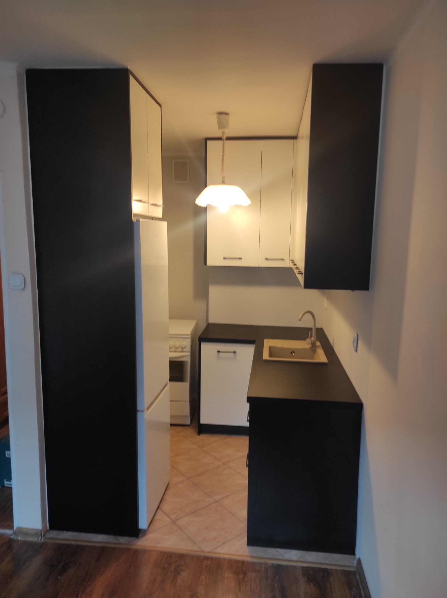 Nowoczesna kuchnia w odcieniach bieli i czerni, z białymi frontami szafek wiszących i czarnymi szafek stojących, z jasnym blatem i zlewozmywakiem, oświetlona wiszącą lampą z kloszem.