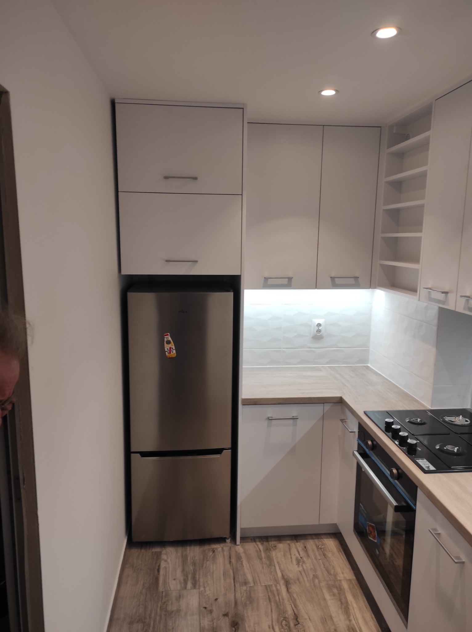 Nowoczesna, jasna kuchnia z białymi szafkami, drewnianym blatem i srebrną lodówką z magnesem w kształcie butelki.