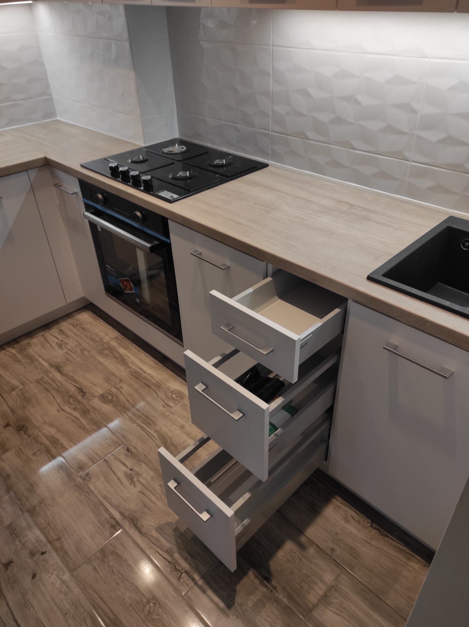 Nowoczesna kuchnia z białymi szafkami, drewnianym blatem i otwartymi szufladami cargo, ukazującymi częściowe wyposażenie. Na blacie płyta grzewcza i czarny zlewozmywak. Podłoga wyłożona panelami...