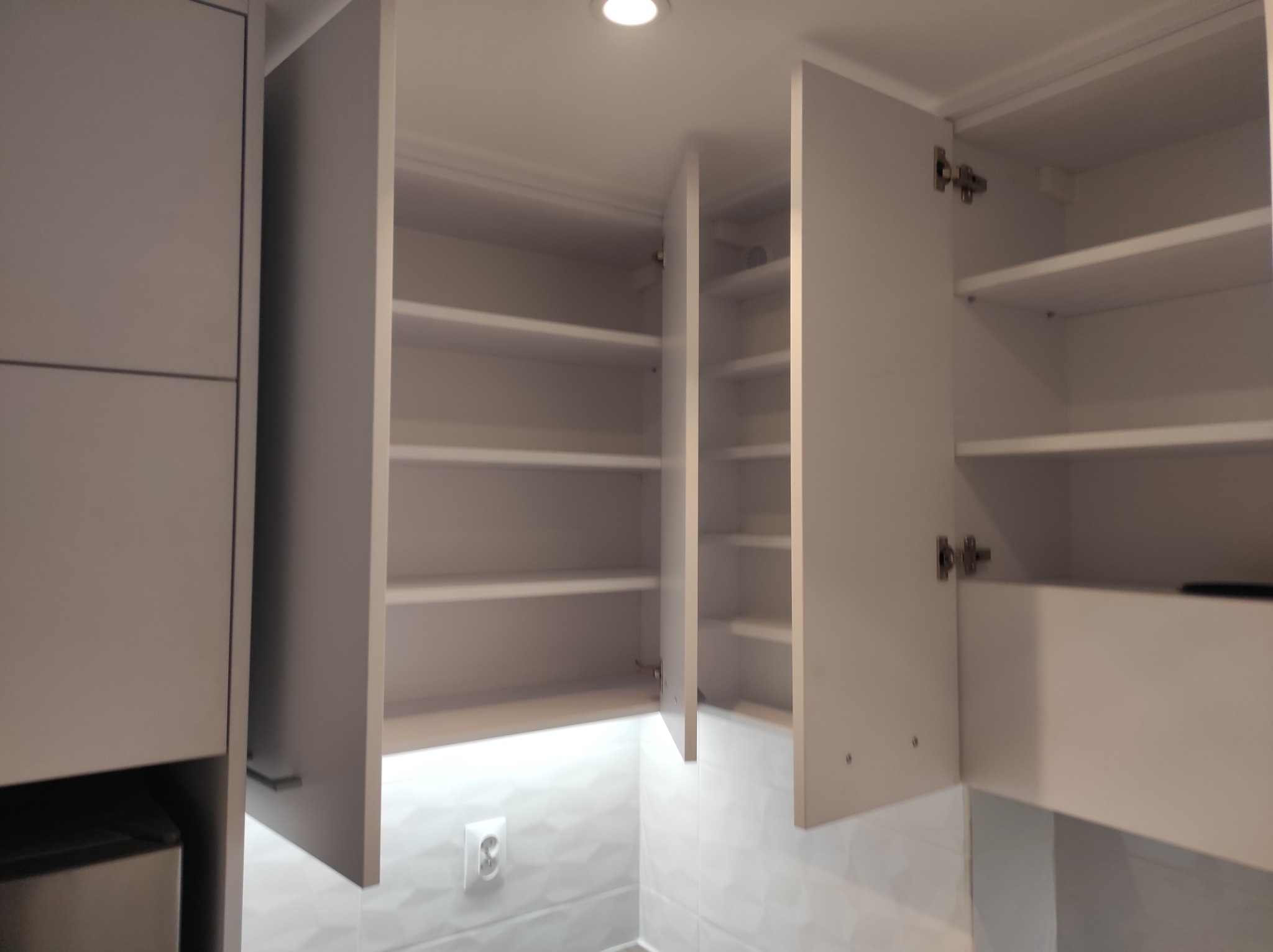 Szare, nowoczesne szafki kuchenne wiszące z otwartymi drzwiczkami, ukazujące puste półki, oświetlone od góry, na tle białych płytek z delikatnym wzorem 3D i gniazdkiem elektrycznym.