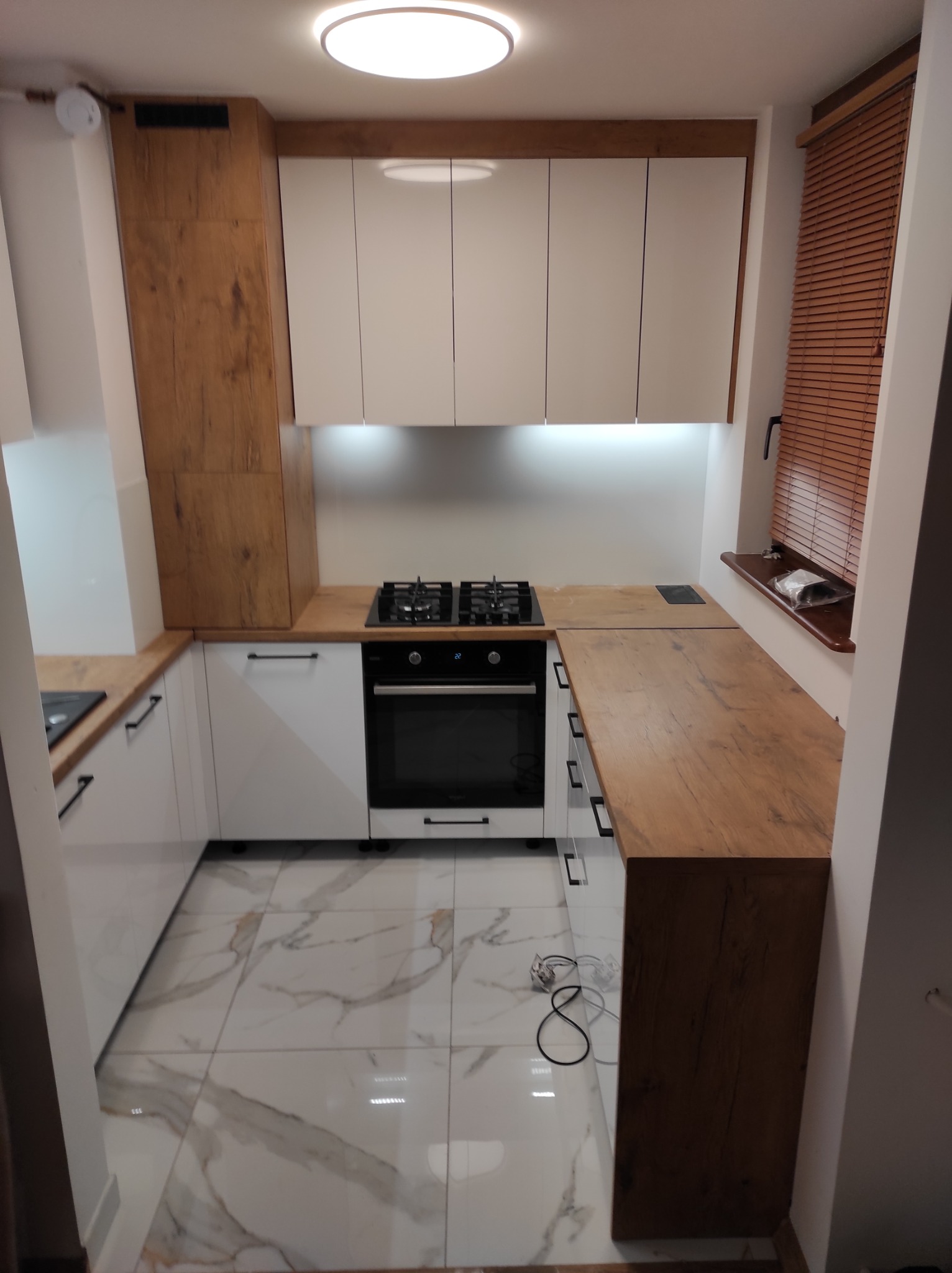 Nowoczesna kuchnia z białymi szafkami i drewnianymi blatami, widok z perspektywy wejścia, marmurowe płytki podłogowe, roleta drewniana w oknie, oświetlenie podszafkowe.