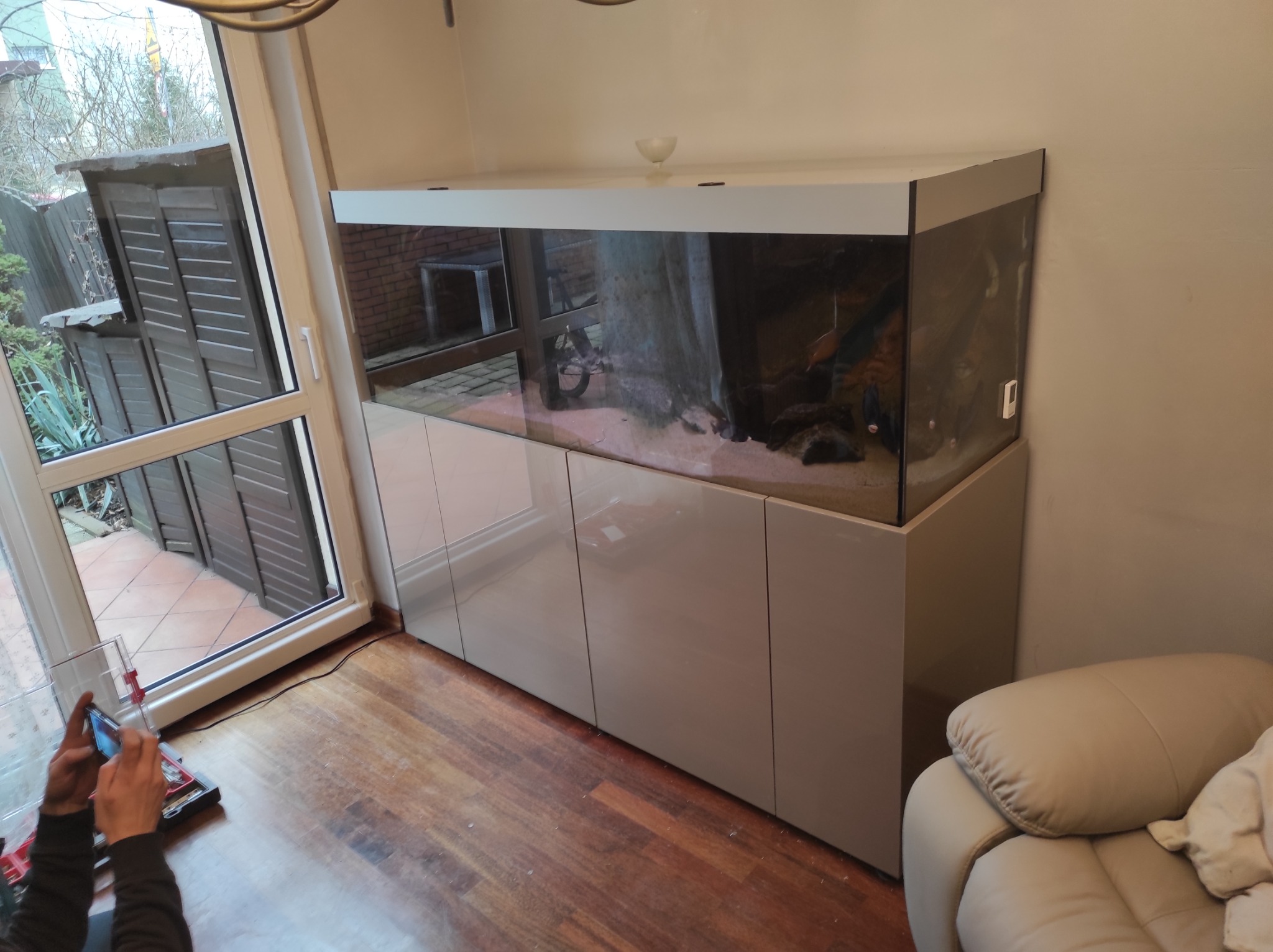 Duże akwarium z szafką w kolorze beżowym, widoczne narzędzia i dłonie osoby montującej, refleksy w szkle akwarium