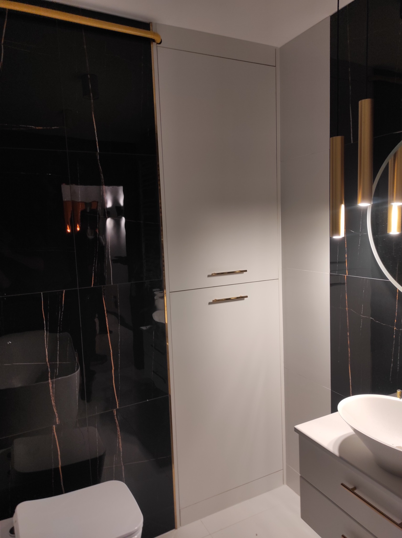 Nowoczesna łazienka z czarnymi płytkami imitującymi marmur z złotymi żyłami, białą szafką z umywalką nablatową, wiszącymi złotymi lampami i wysoką, wąską, białą szafką.