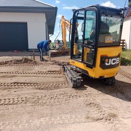 Alexbruk - Żółta mini koparka JCB na piaszczystym terenie z widocznymi śladami gąsienic, pracownik w niebieskiej bluzie z łopatą obok wykopu, w tle dom z szarą bramą garażową i fragment drugiego domu.