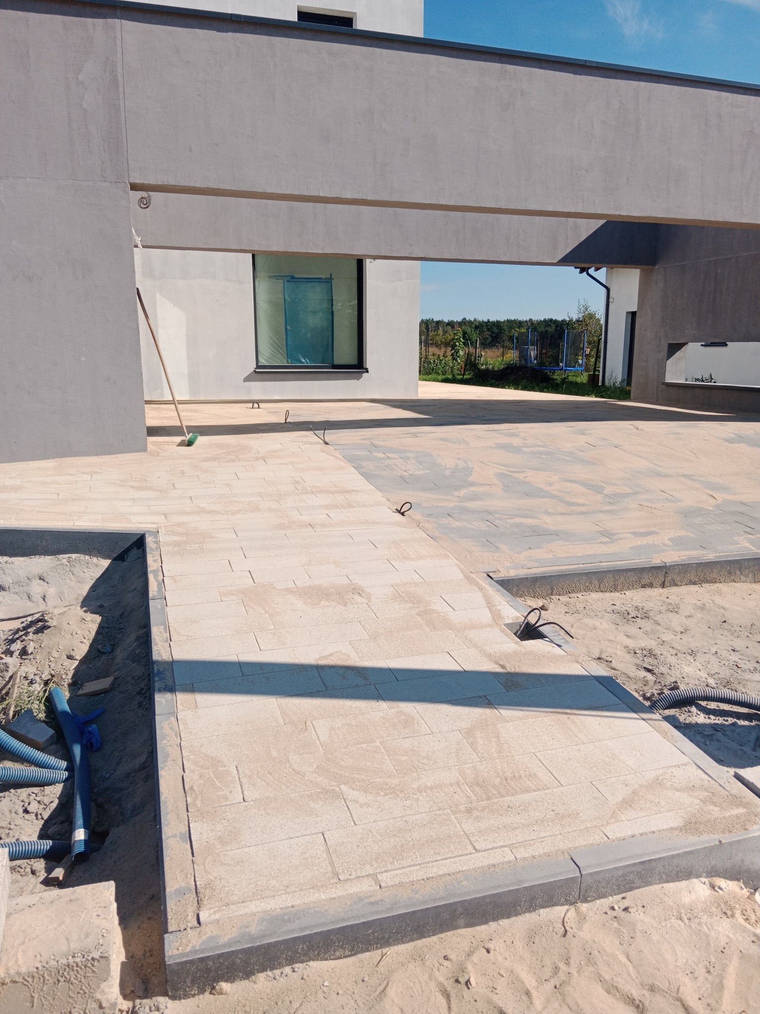 Fragment nowej nawierzchni z jasnej kostki brukowej na posesji, widoczne kable i węże, piasek i fragment budynku w tle.