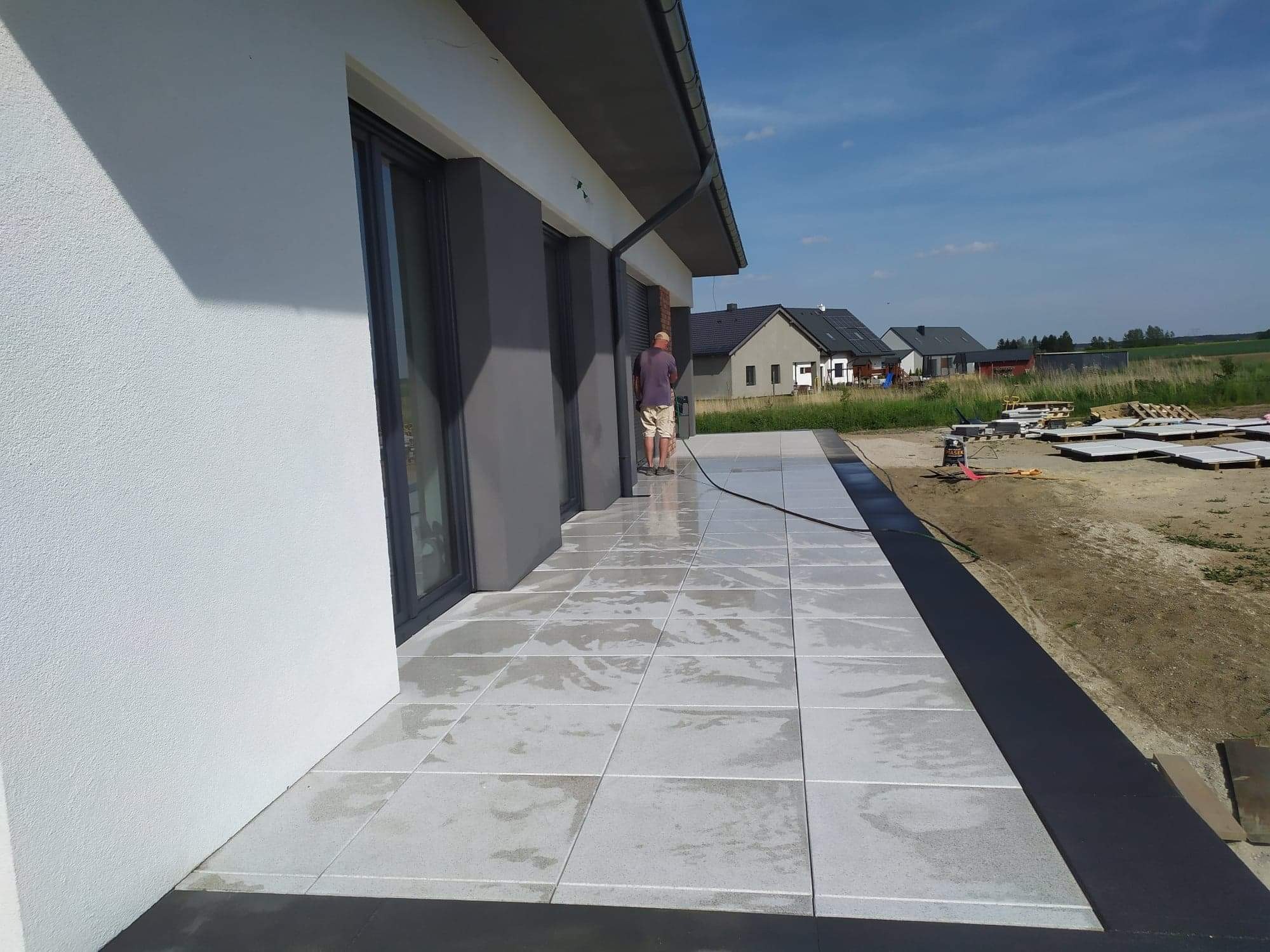 Świeżo ułożony taras z jasnoszarych płyt, myty pod ciśnieniem przez mężczyznę w krótkich spodenkach, w tle widoczne domy i palety z kostką.