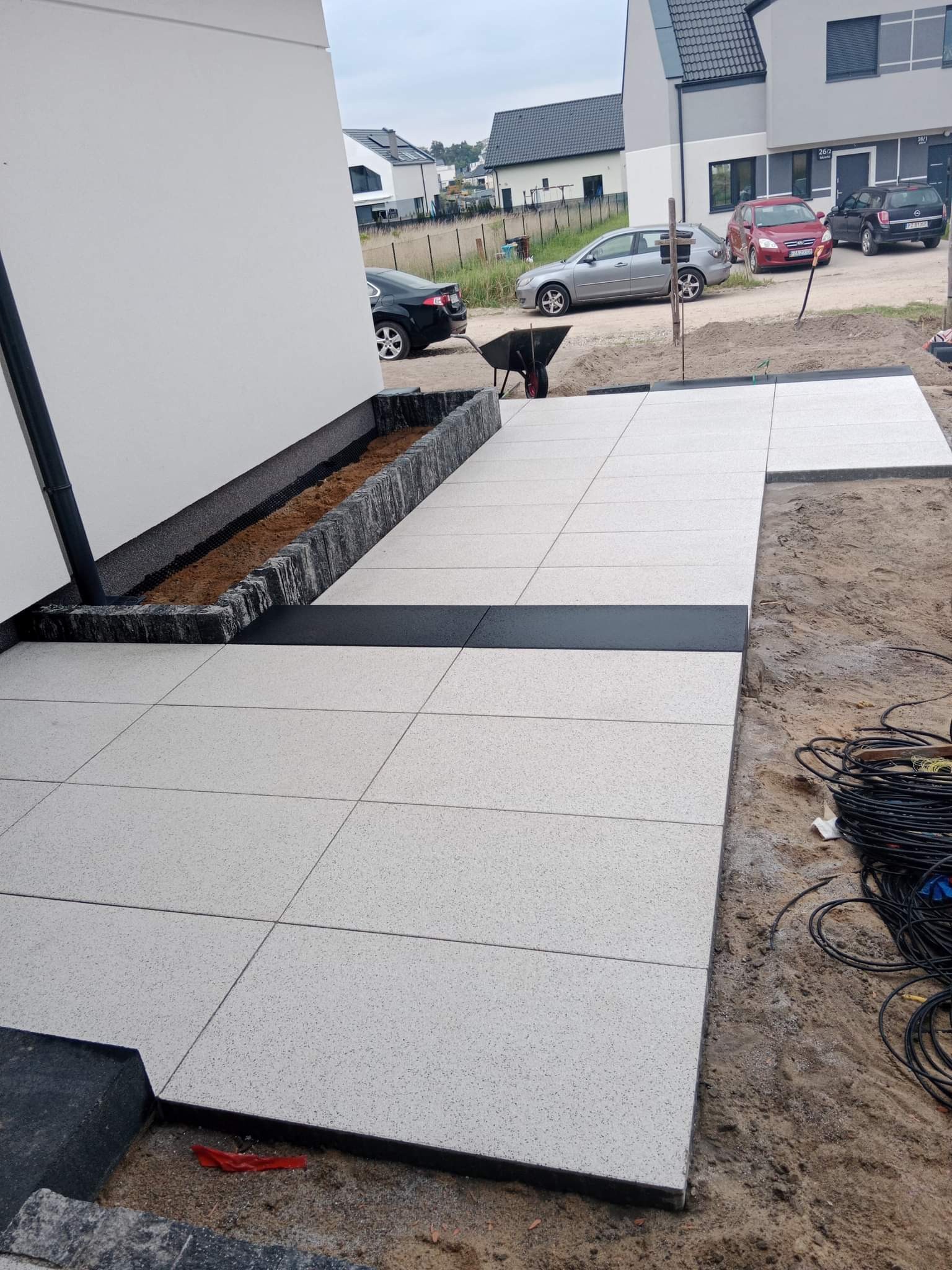 Ułożona, jasnoszara kostka brukowa z ciemnymi paskami oddzielającymi, obok ściany budynku i rabaty z ziemią, w tle zaparkowane samochody i budynki.