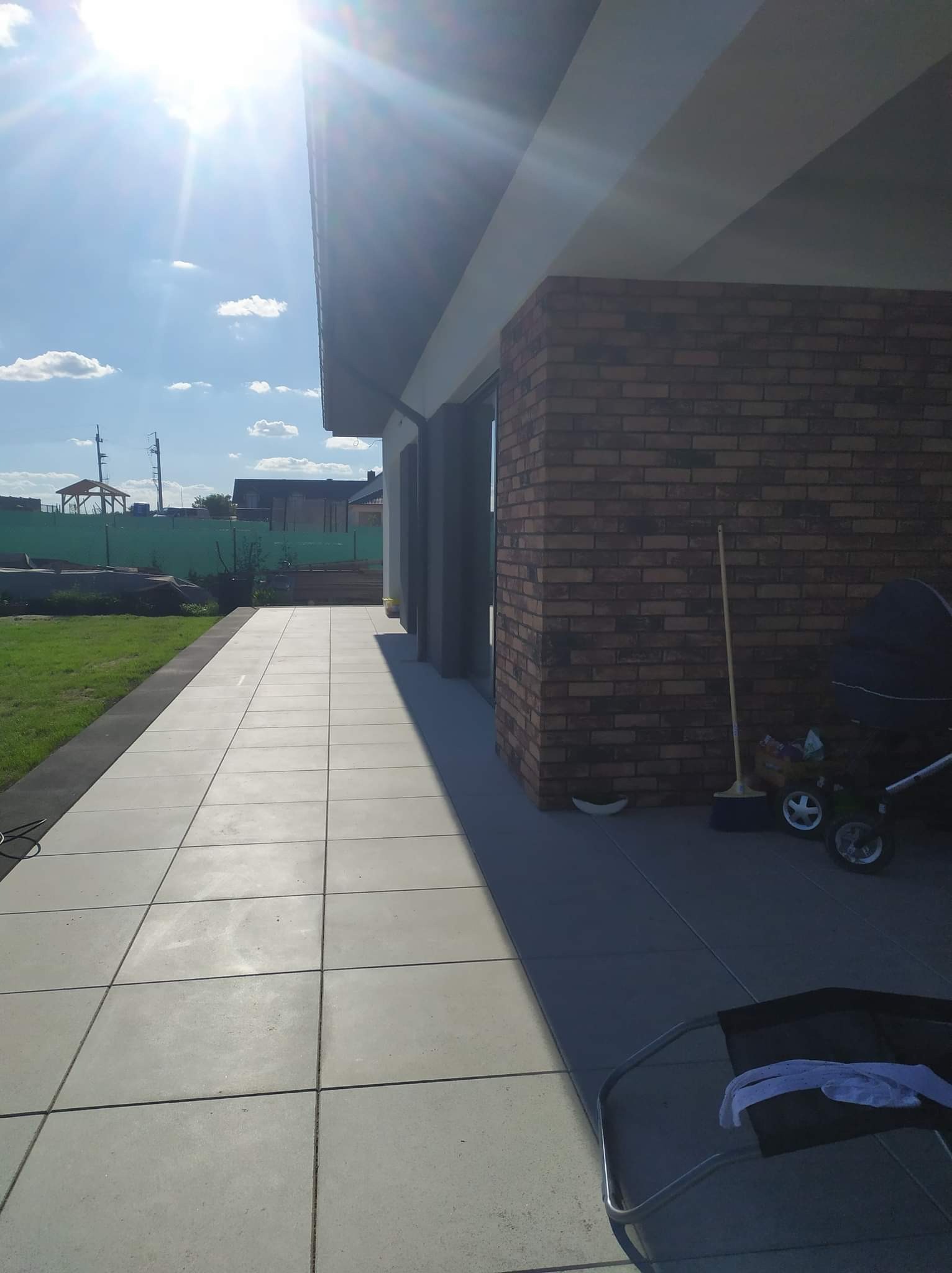 Ułożona nawierzchnia z szarych płyt betonowych przed nowoczesnym domem z ceglaną elewacją, widoczne narzędzia i wózek dziecięcy.