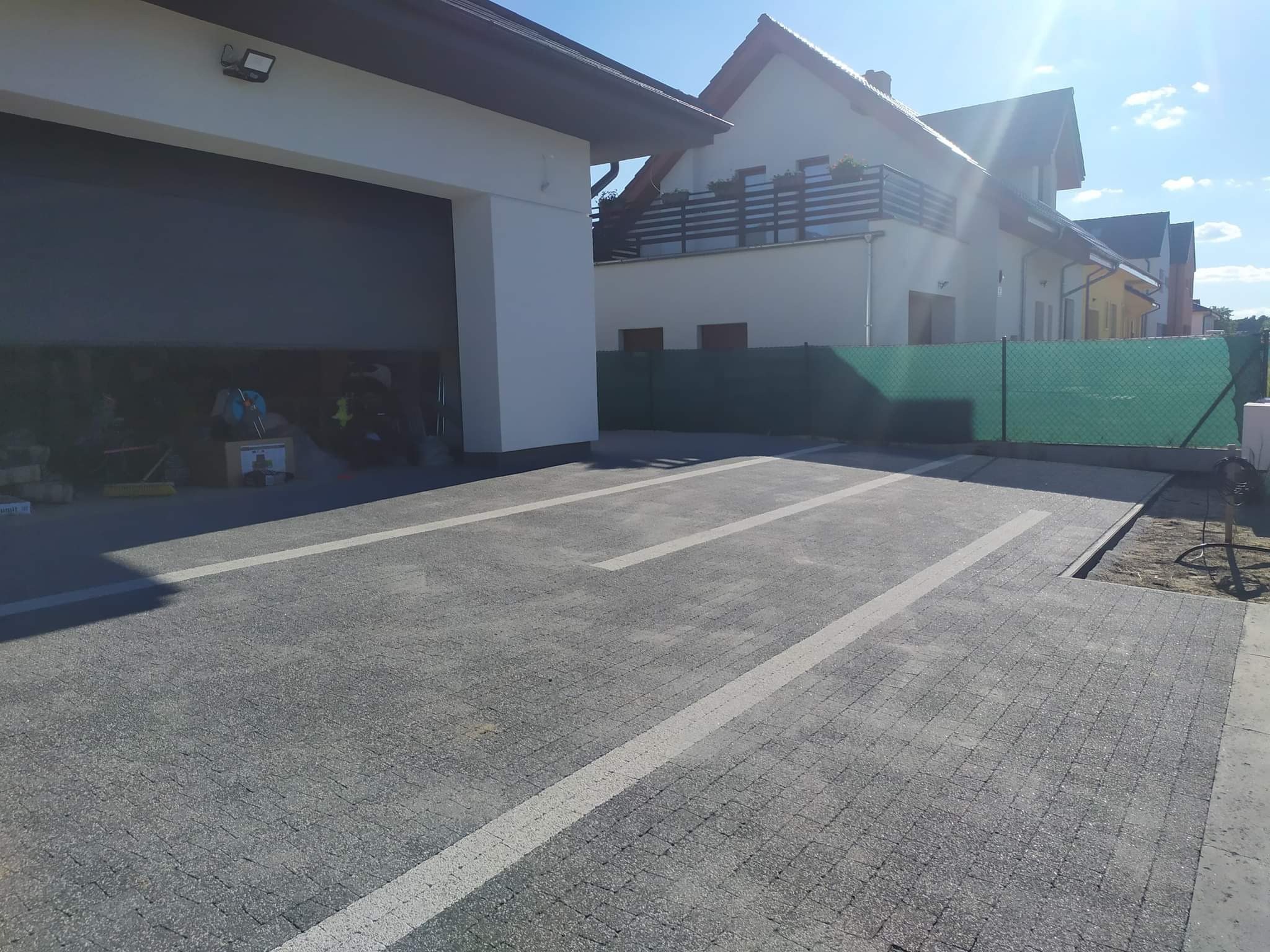 Szara kostka brukowa z białymi liniami wyznaczającymi miejsca parkingowe, przed budynkiem z otwartym garażem i domem jednorodzinnym w tle.