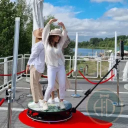 Kobieta w białym stroju i kapeluszu tańcząca na obrotowej platformie fotobudki 360 na tle jeziora i błękitnego nieba.