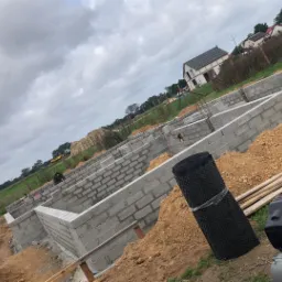 Wykopane fundamenty domu z bloczków betonowych, z widocznym zbrojeniem i izolacją przeciwwilgociową w rolkach, obok kompresor.