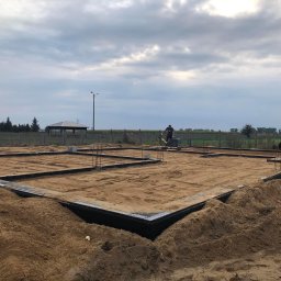 SIKOR-BUD - Przygotowanie fundamentów pod budowę domu jednorodzinnego: widoczny zarys fundamentów, piasek zasypany wewnątrz, pracownik ubijający ziemię, w tle krajobraz z altaną i pochmurnym niebem.