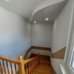 SIKOR-BUD - Klatka schodowa z drewnianymi schodami i balustradą, ściany pomalowane na biało z dekoracyjnym pasem w kolorze brązowym, sufit z podwieszanym elementem i oświetleniem punktowym.