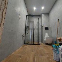 SIKOR-BUD - Nowoczesna łazienka z szarymi płytkami ściennymi, drewnopodobną podłogą, kabiną prysznicową z czarnymi ramami i wiszącą toaletą. Częściowo wykończona ściana po lewej stronie sugeruje trwający remont.