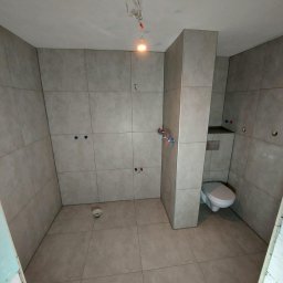 SIKOR-BUD - Łazienka w trakcie wykańczania: ułożone płytki ścienne i podłogowe w odcieniach szarości, widoczne instalacje wodne, podwieszana toaleta, surowe oświetlenie budowlane.