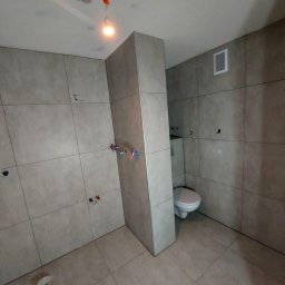 SIKOR-BUD - Wnętrze łazienki w trakcie remontu, widoczne szare płytki na ścianach i podłodze, częściowo zamontowana toaleta, wystające przewody elektryczne i niedokończone przyłącza wodne.