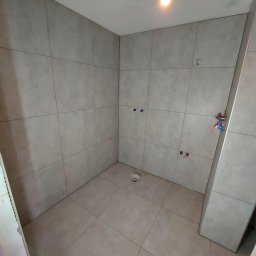 SIKOR-BUD - Wnętrze łazienki w trakcie remontu, widoczne płytki na ścianach i podłodze, instalacja wodna i elektryczna przygotowana, kubek na podłodze.