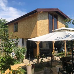 SIKOR-BUD - Dom w trakcie ocieplania, ściany pokryte płytami OSB, widoczne okna z brązowymi ramami, w tle błękitne niebo i zieleń drzew, w pierwszym planie altana ogrodowa z białym dachem.