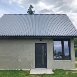 SIKOR-BUD - Niewykończony budynek z szarą blachodachówką, szarą elewacją przed położeniem tynku, ciemnymi drzwiami z pionowym przeszkleniem i oknem z widocznym odbiciem drzewa.