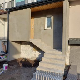 SIKOR-BUD - Nowe, betonowe schody prowadzące do wejścia budynku, ściany w trakcie szpachlowania, widoczny cień osoby fotografującej.