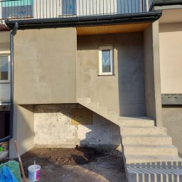 SIKOR-BUD - Narożnik budynku po szpachlowaniu, widoczne surowe, betonowe schody wejściowe, fragment elewacji z oknem i skrzynką gazową, narzędzia i materiały budowlane w tle.