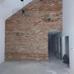 SIKOR-BUD - Surowa ceglana ściana z trzema nowoczesnymi lampami wiszącymi w przestronnym, jasnym wnętrzu z surową podłogą i otwartym przejściem do korytarza.