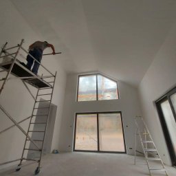 SIKOR-BUD - Malowanie sufitu w wysokim pomieszczeniu z dużymi oknami zabezpieczonymi folią, pracownik na rusztowaniu używa wałka, widoczna drabina. Sufit częściowo pomalowany.