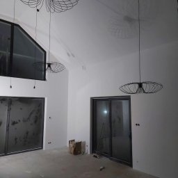 SIKOR-BUD - Wnętrze w trakcie wykańczania z trzema nowoczesnymi lampami wiszącymi o ażurowych kloszach, rzucającymi cienie na gładką, białą ścianę, widoczne surowe podłoże i okna z ciemnymi ramami.