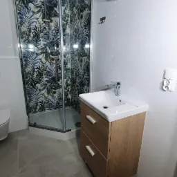 Nowoczesna łazienka z kabiną prysznicową w tropikalne wzory, umywalką z szafką z jasnego drewna i geometrycznymi wieszakami na białych płytkach.