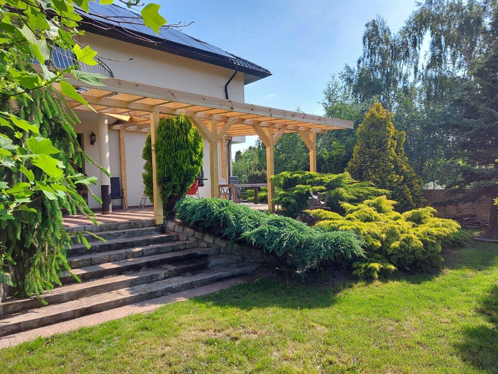 Drewniana pergola z jasnego drewna, częściowo zadaszona, przylegająca do budynku z panelami słonecznymi, widok z poziomu trawnika, z kamiennymi schodami i ozdobnymi krzewami iglastymi na pierwszym...