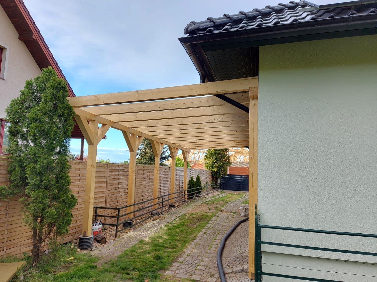 Drewniana konstrukcja zadaszenia tarasu z widocznymi elementami montażowymi, przylegająca do jasnej ściany budynku, z tujami i ogrodzeniem w tle.