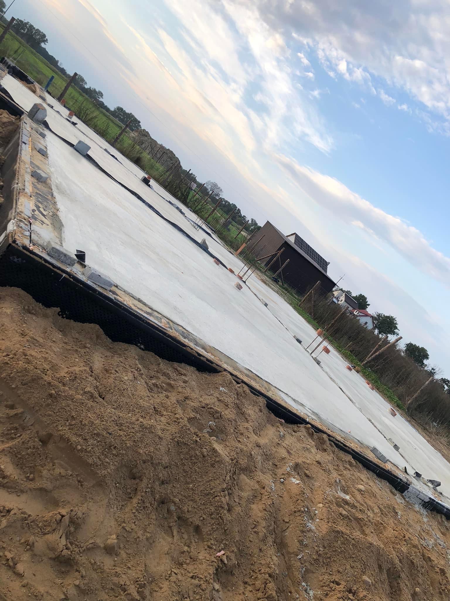 Betonowa płyta fundamentowa z izolacją pionową i poziomą, przygotowana pod budowę domu jednorodzinnego na tle krajobrazu wiejskiego.