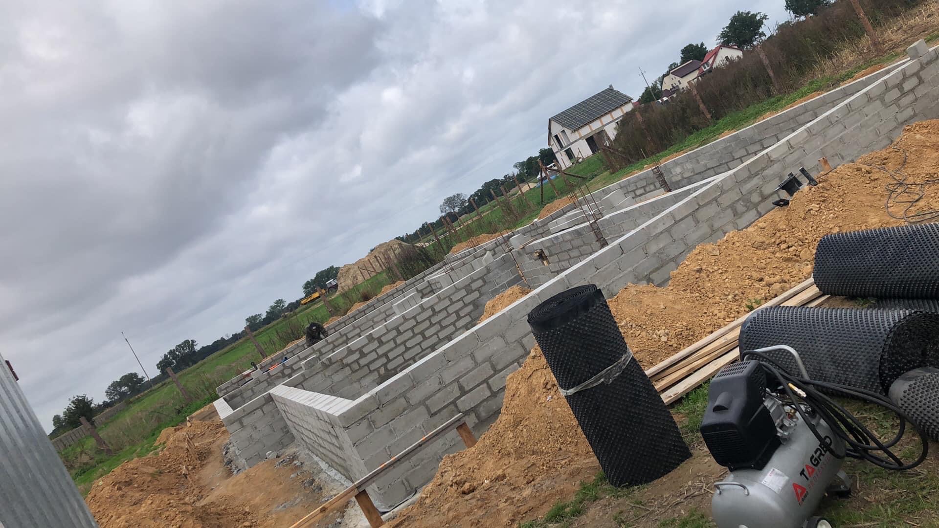 Wykopane fundamenty domu z bloczków betonowych, z widocznym zbrojeniem i izolacją przeciwwilgociową w rolkach, obok kompresor.