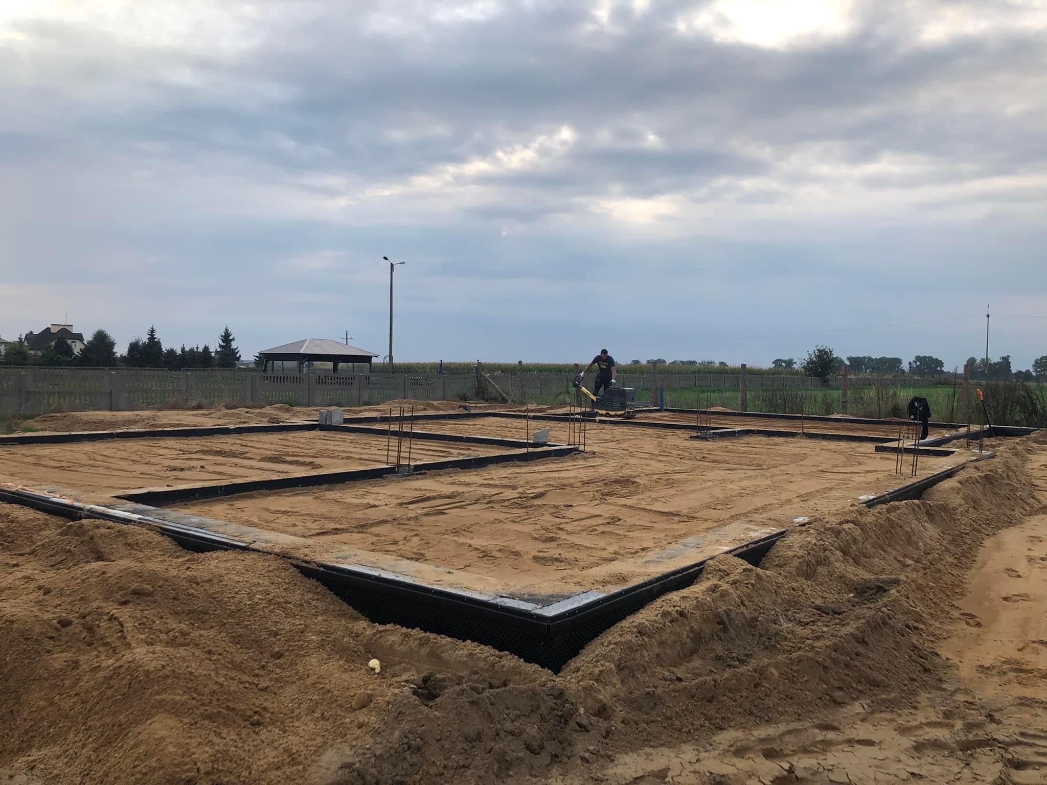 Przygotowanie fundamentów pod budowę domu jednorodzinnego: widoczny zarys fundamentów, piasek zasypany wewnątrz, pracownik ubijający ziemię, w tle krajobraz z altaną i pochmurnym niebem.