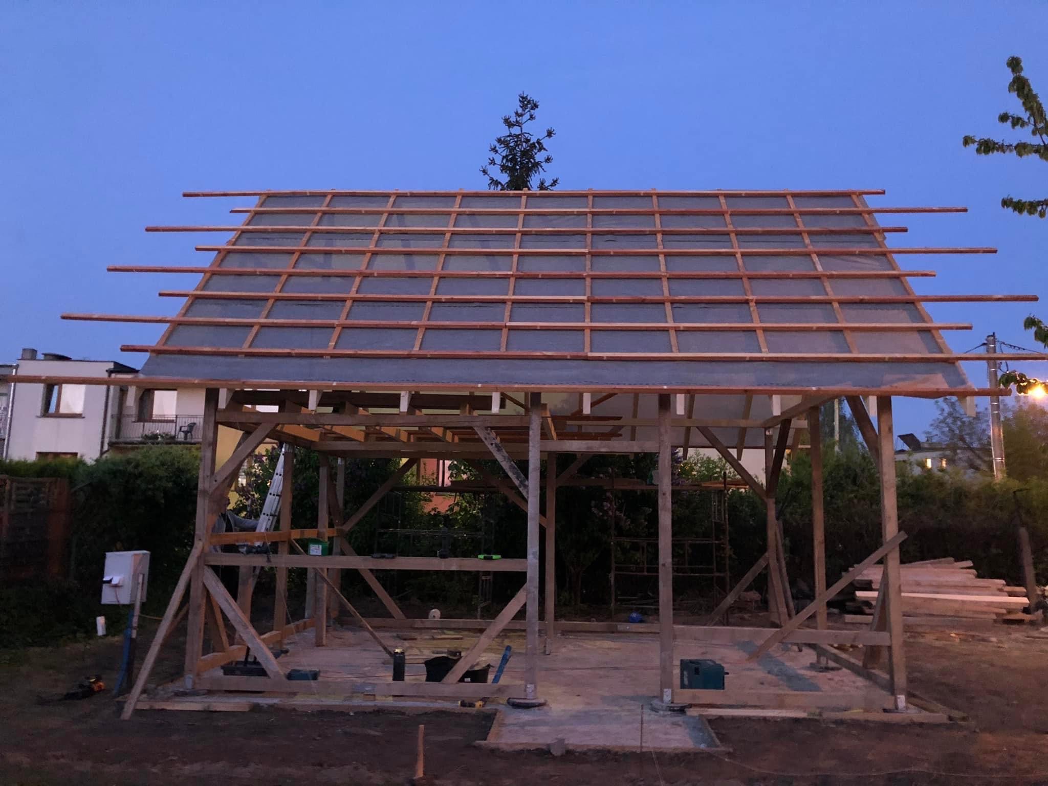 Drewniana konstrukcja z częściowo wykonanym dachem, widoczne deski i folia, w tle zabudowa jednorodzinna i zieleń, wieczorny krajobraz.