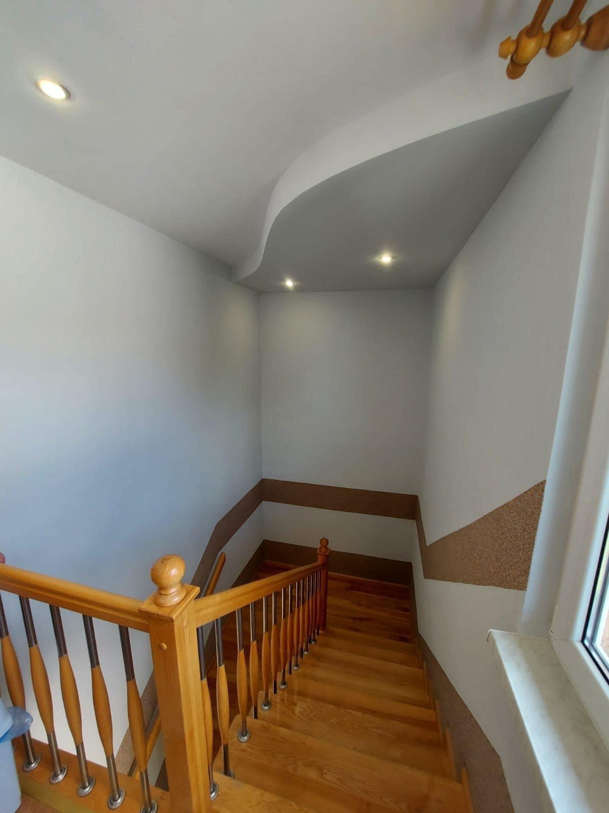 Klatka schodowa z drewnianymi schodami i balustradą, ściany pomalowane na biało z dekoracyjnym pasem w kolorze brązowym, sufit z podwieszanym elementem i oświetleniem punktowym.
