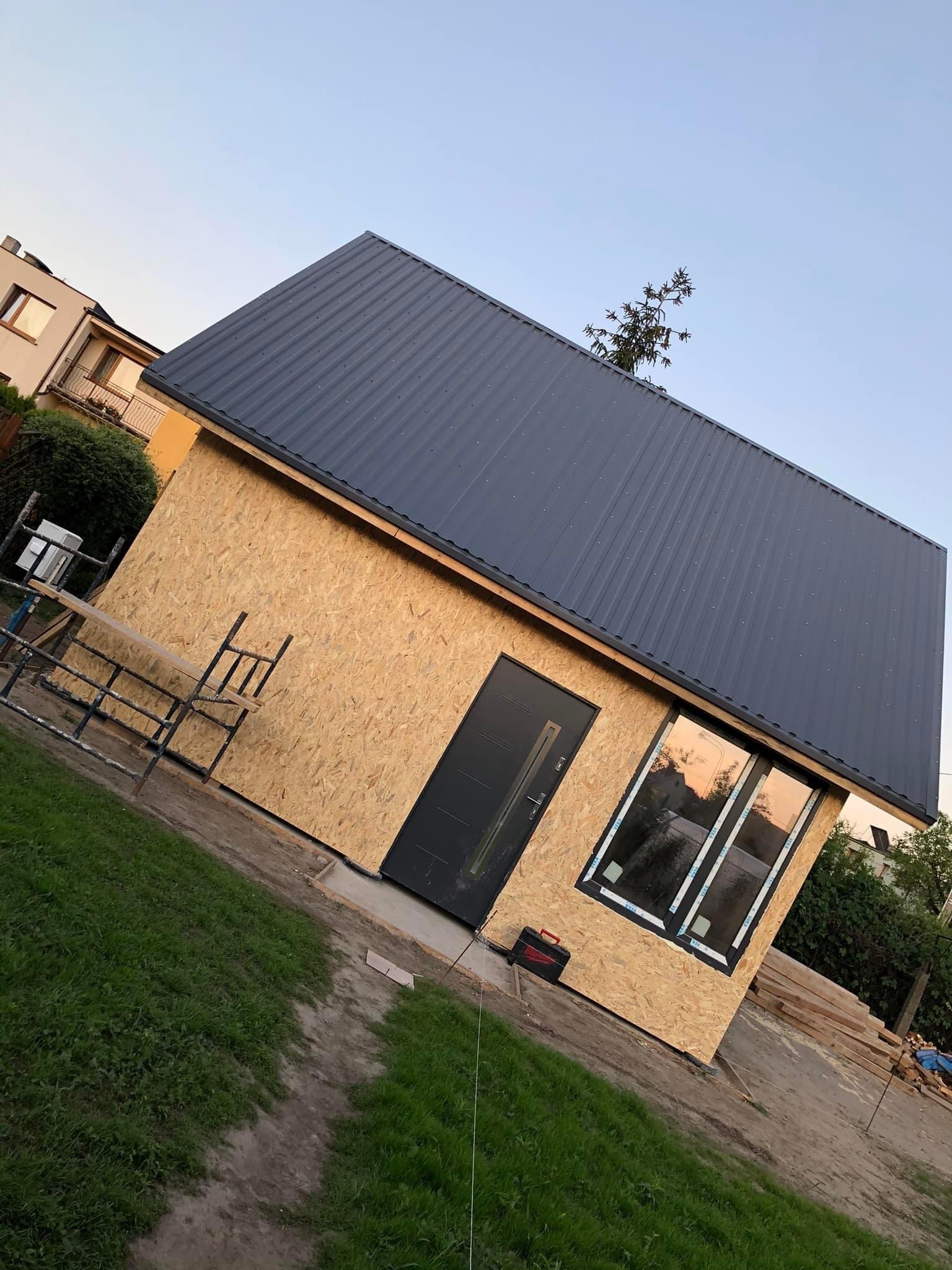 Niedokończony budynek gospodarczy z dachem z blachy trapezowej, ścianami z płyt OSB, wstawionymi oknami i drzwiami, widoczne rusztowanie i narzędzia.