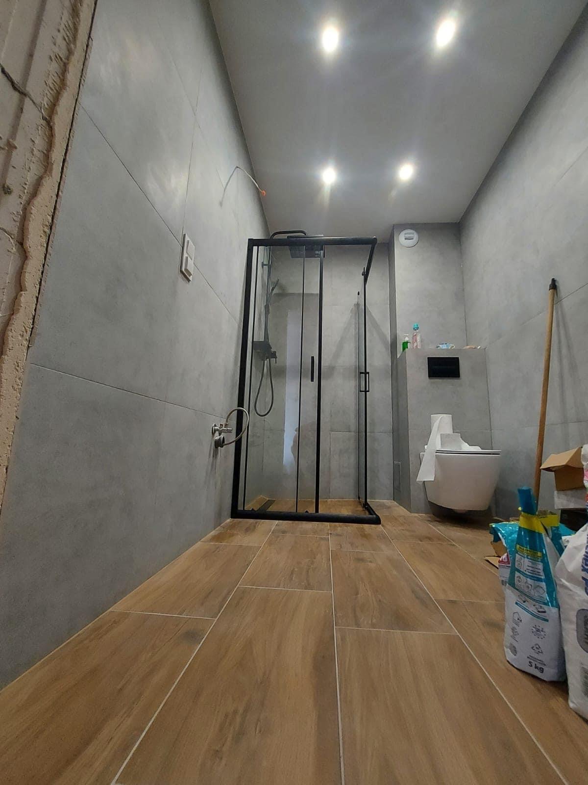 Nowoczesna łazienka z szarymi płytkami ściennymi, drewnopodobną podłogą, kabiną prysznicową z czarnymi ramami i wiszącą toaletą. Częściowo wykończona ściana po lewej stronie sugeruje trwający remont.